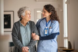 A young nurse linked arms with an older woman with a walking stick in a care home.