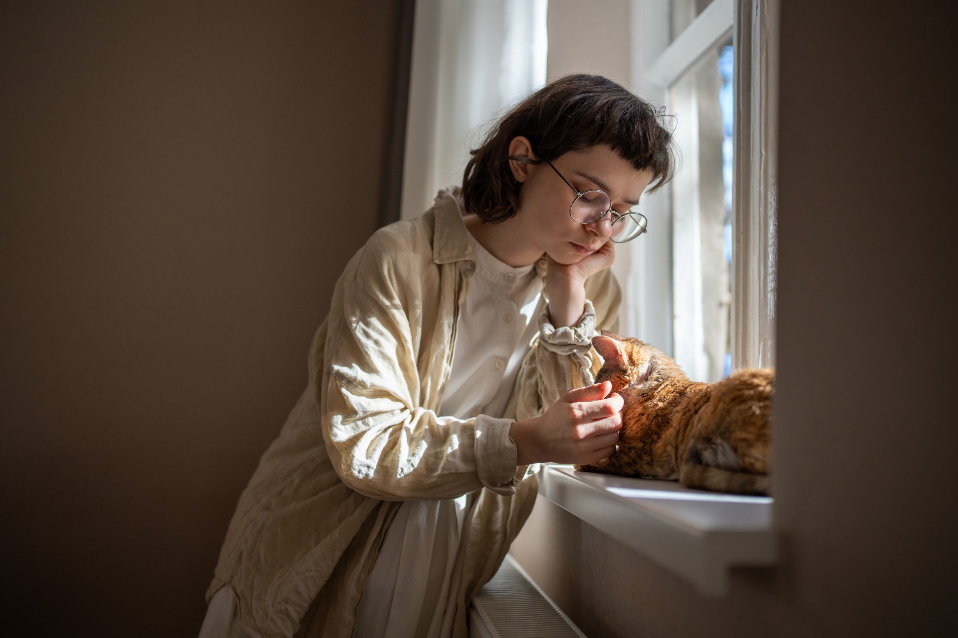 A lonely young woman petting her cat on the window sill.