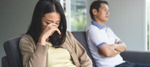 a couple facing away from each other on a sofa. the woman is crying while the man has his arms folded