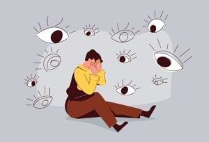 A sad man sat on the ground with lots of large eyes watching him.