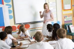 A female teacher at the front of class teaching a table full of children.
