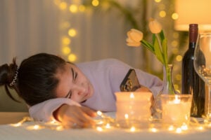 A single girl lay with her head on the table looking at her phone. Candles and a bottle of wine on the table.