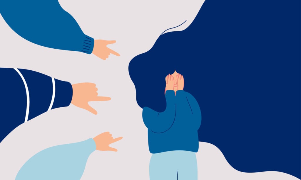 Depressed girl cries and covers her face with her hands surrounded by the hands of her peers pointing at her.