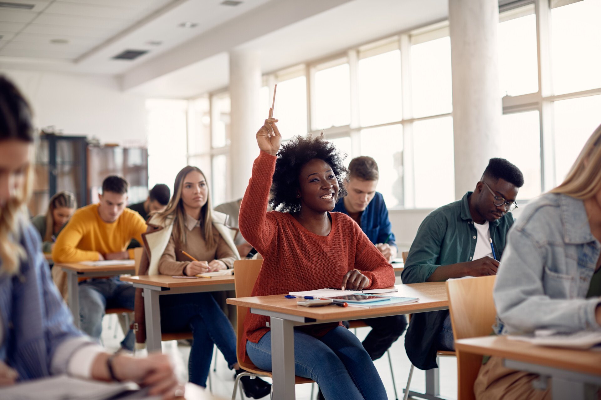 A group of college students in a classroom with one student raising her hand to ask a question