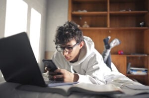A teenage boy using his smart phone while lay on bed with laptop.