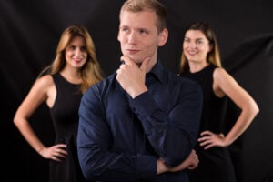 A confident man doing a thinking pose and smirking while there are two smiling women looking at him in the background.